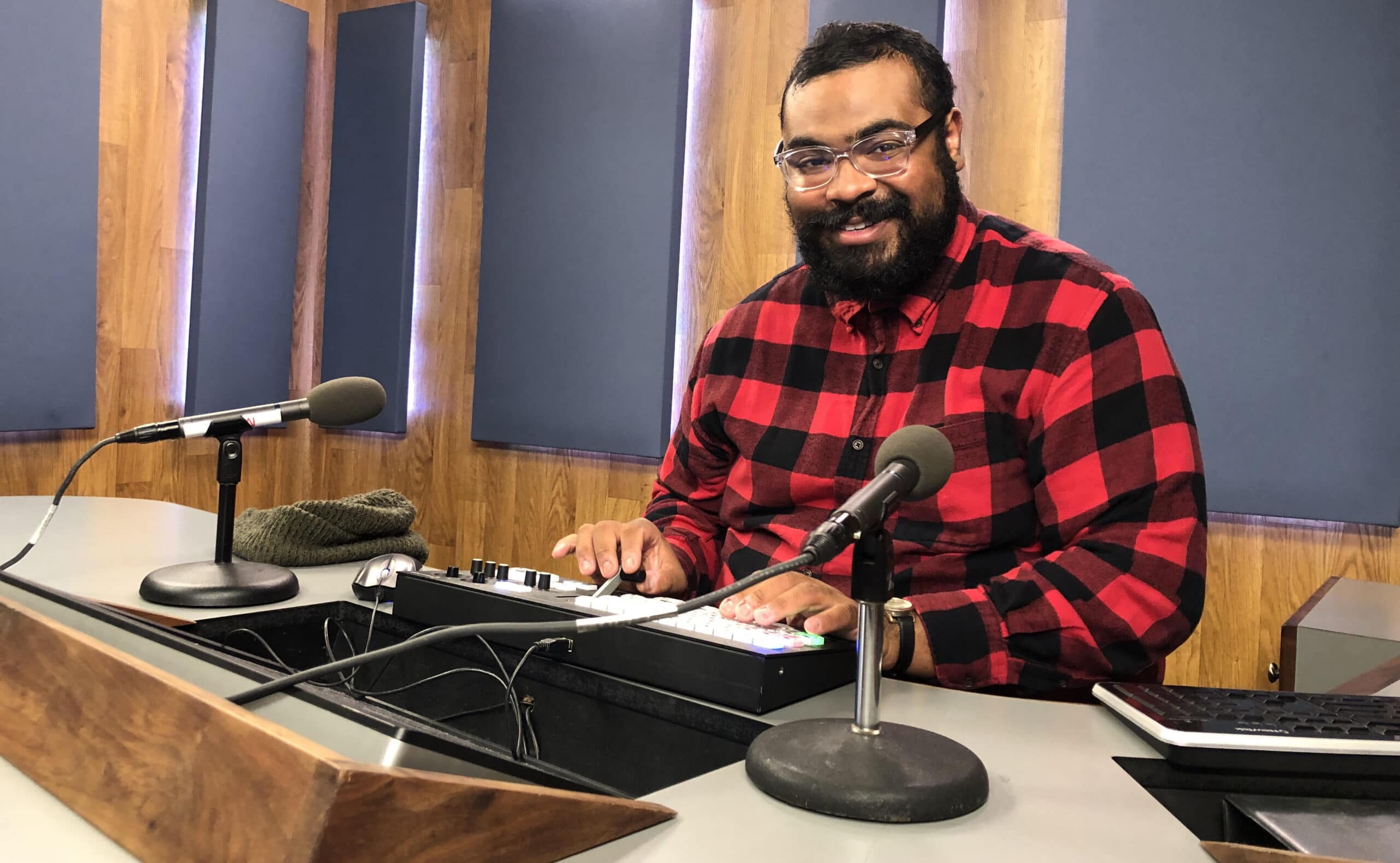 Image of a smiling Black man in a red & black check shirt sitting behind a podcast / audio recording set up.