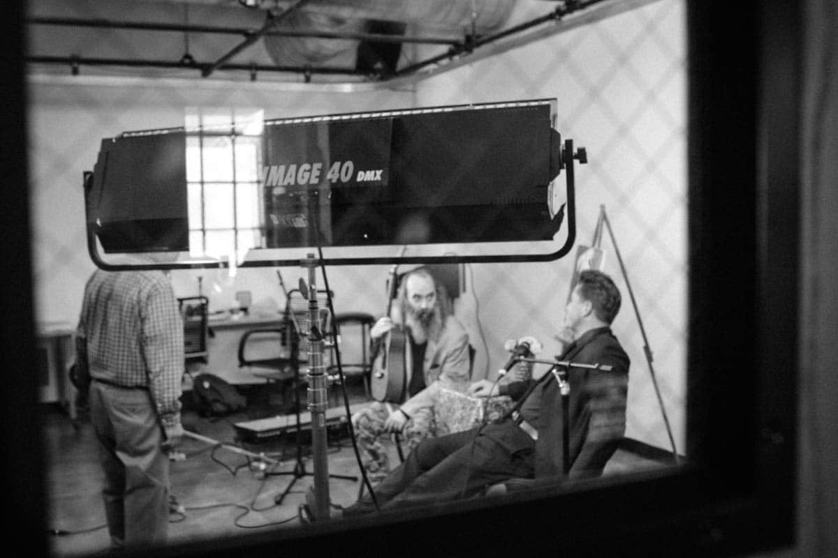 Black and white behind-the-scenes photo taken through a window in a door of group of men filming a scene in a TV studio.
