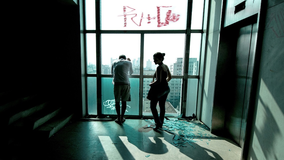 Image of a man and a woman silhouetted in front of a window with gray-blue cityscape beyond,
