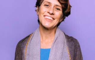 Kia smiles broadly, head tilted, short wavy hair, turquoise shirt, grey wrap, lavender background