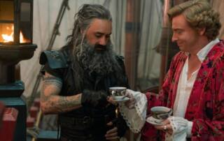 Bearded pirate Blackbeard and aristocrat Stede share tea on a pirate ship