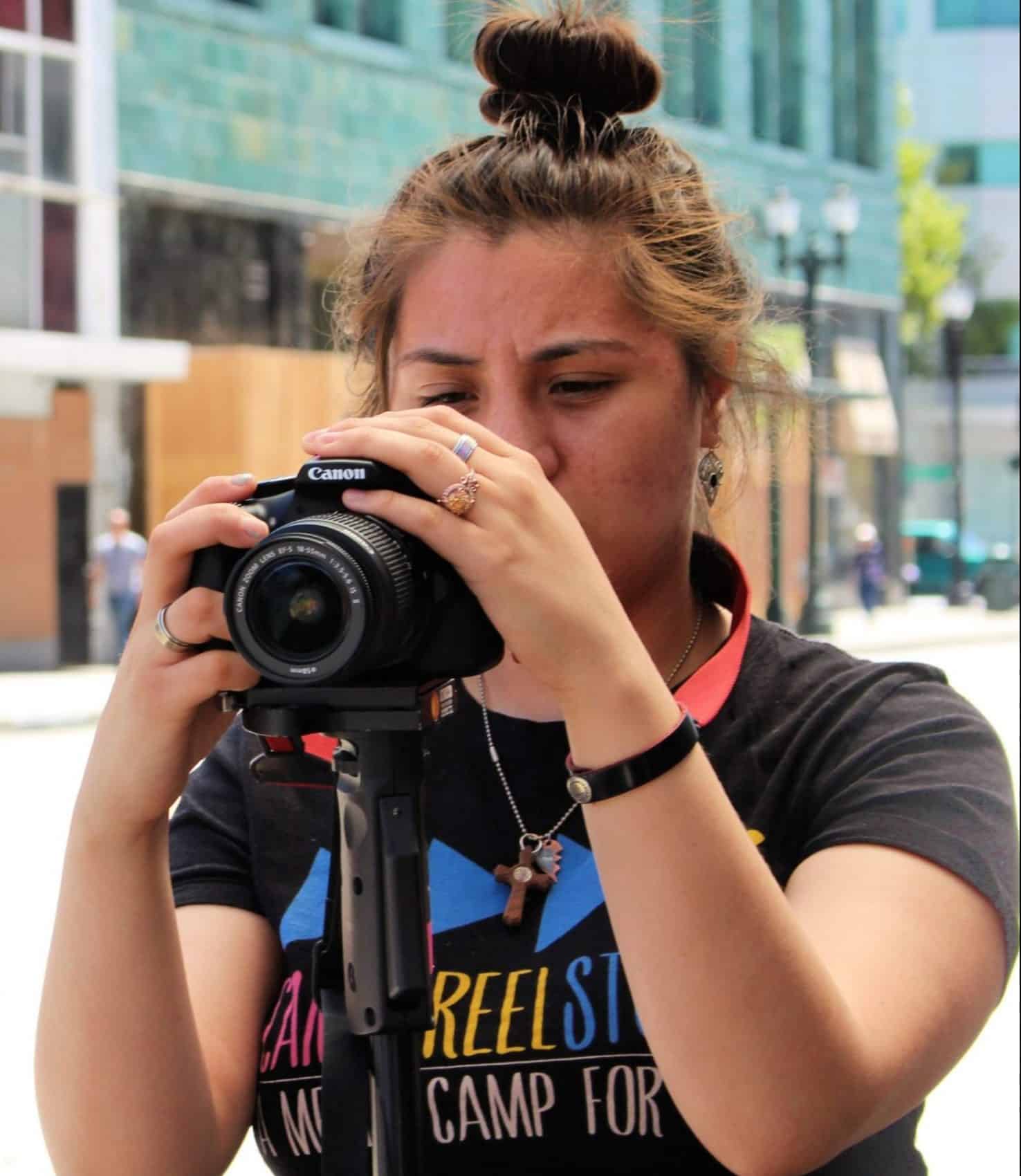 Liz Tril, a young woman with brown hair up in a bun, looks through the lens of a digital camera.